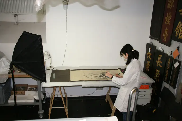 A person wearing a mask and a white lab coat works on a large piece of fabric at a table. The environment includes hanging artworks and a camera setup nearby, suggesting an art preservation or documentation process.