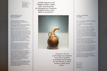 A ceramic pitcher with a curved handle and decorative patterns is displayed in a museum setting. Two text panels flanking the pitcher feature German and English inscriptions discussing the relationship between ancient cultures, indigenous peoples, and ecological processes.