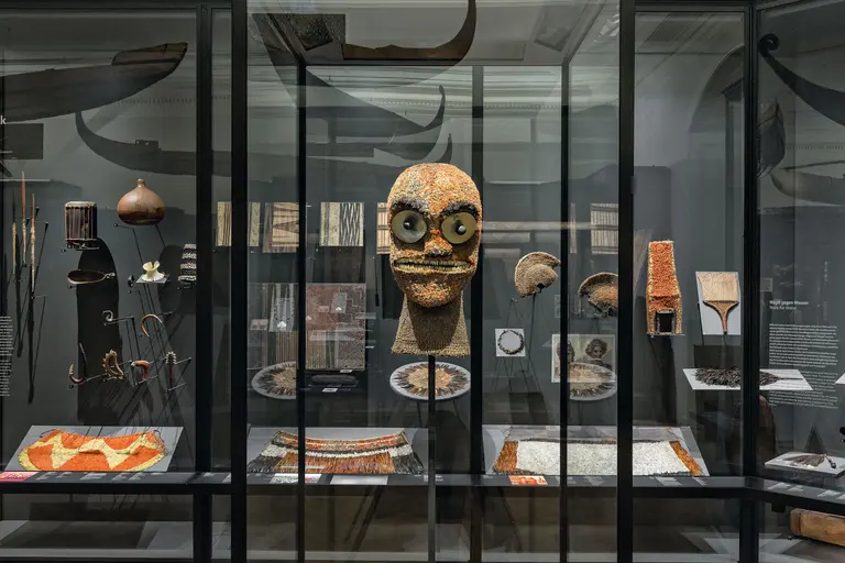 Eine Vitrine in einem Museum zeigt ethnographische Objekte, darunter eine auffällige Maske mit großen Augen und orangefarbener Oberfläche. Um die Maske herum sind weitere Gegenstände wie Werkzeuge, Textilien und kunsthandwerkliche Erzeugnisse angeordnet.
