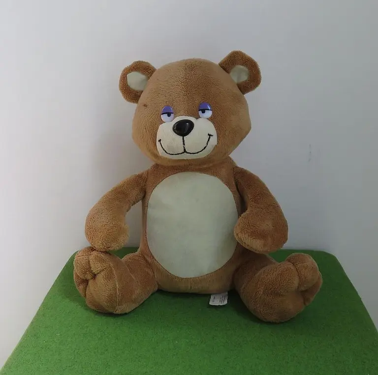 Ein kuscheliger, brauner Teddy sitzt auf einem grünen Untergrund. Er hat eine runde Form, einen breiten Mund und einen freundlichen Gesichtsausdruck, mit leicht geschlossenen Augen, als ob er schläfrig ist. Seine Ohren sind rund und er hat eine helle Bauchpartie.