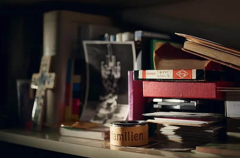 Auf einem Tisch sind mehrere gestapelte Bücher, Zeitschriften und ein Fotodruck zu sehen. Ein kleines Häufchen mit der Aufschrift „familien“ ragt hervor. Der Hintergrund ist unscharf, und das Licht fällt sanft auf die Gegenstände.