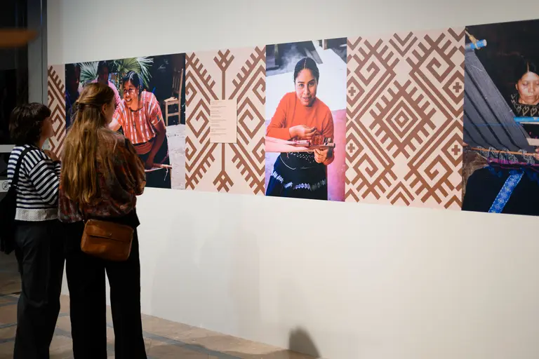 Zwei Frauen betrachten eine Bilderwand mit Fotografien von Frauen, die traditionelle Handwerkskünste ausüben. Die Bilder sind in einem bunten, ethnischen Muster angeordnet. Im Hintergrund sind verschiedene Muster und Farben zu sehen, die die kulturelle Bedeutung der Kunstwerke unterstreichen.