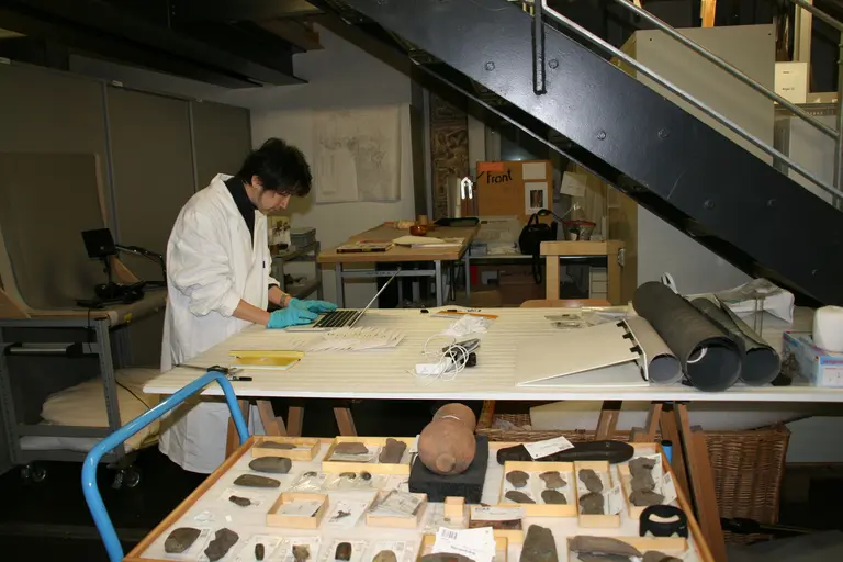 A researcher in a white lab coat and blue gloves examines artifacts on a table in a laboratory setting. Various stone tools and items are displayed in wooden trays. The environment includes shelves and a staircase, indicating a workspace for handling historical materials.