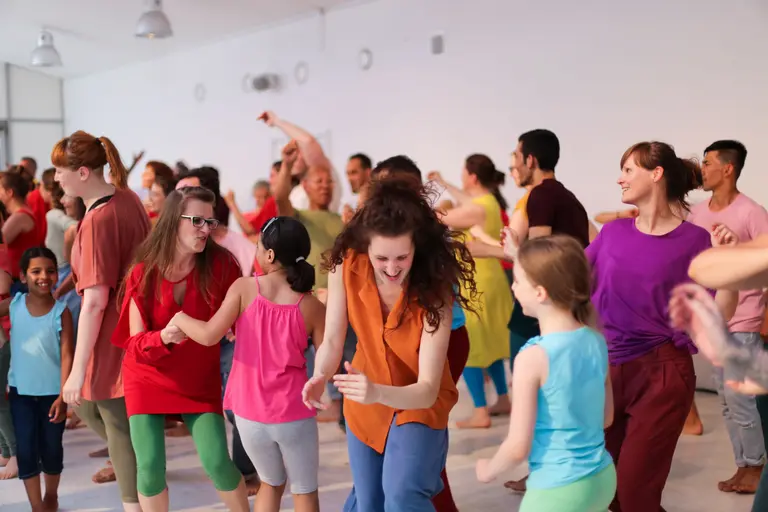 Eine große Gruppe von Menschen tanzt und bewegt sich fröhlich in einem hellen Raum. Die Teilnehmer tragen farbenfrohe Kleidung und scheinen Spaß zu haben. Die Atmosphäre ist lebhaft und dynamisch.