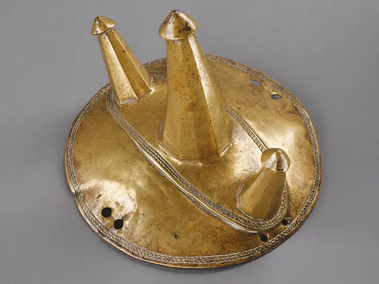 Eine runde, flache goldene Schale mit drei kegelförmigen Erhebungen. Die Kegel haben spitze Spitzen und sind unterschiedlich hoch. Auf der Oberfläche sind feine Verzierungen zu erkennen. Der Hintergrund ist grau.