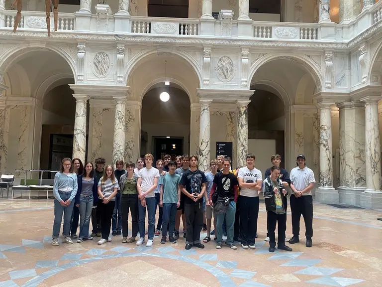 Eine Gruppe von etwa 20 Jugendlichen steht in einem stilvollen Innenraum mit hohen Säulen und marmorartigen Wänden. Sie posieren zusammen, einige lächeln, während andere neutral schauen. Im Hintergrund ist ein großer, heller Raum mit Sitzgelegenheiten sichtbar.