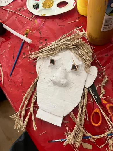 A handmade cardboard face with straw for hair and cut-out eyes is placed on a red table covered in art supplies. Scissors and colorful paints are nearby, indicating an art project in progress.