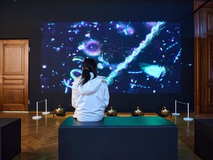 Eine Person sitzt auf einer Bank und schaut auf eine große Projektion an der Wand, die bunte, abstrakte Muster und Bewegungen zeigt. Im Vordergrund sind einige kleine Objekte platziert, und die Wände des Raumes sind dunkel.