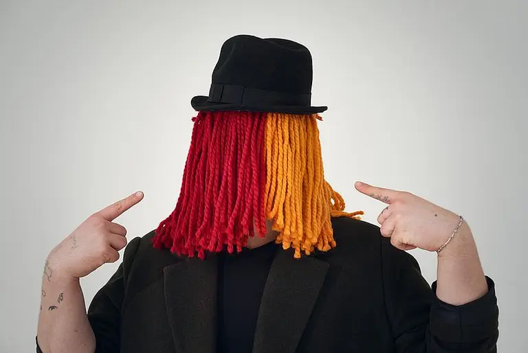 Eine Person mit einer auffälligen Perücke in Rot und Gelb trägt einen schwarzen Hut. Sie zeigt mit beiden Händen auf sich selbst. Der Hintergrund ist neutral und hell, was den Fokus auf die Person lenkt.