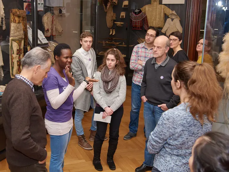 Eine Gruppe von Menschen steht in einem Museum und hört einer Führerin zu, die Informationen über Exponate teilt. Im Hintergrund sind traditionelle Kleidungsstücke und Artefakte zu sehen. Die Teilnehmer scheinen interessiert und engagiert.