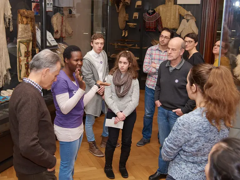 Eine Gruppe von Menschen steht in einem Museum und hört einer Führerin zu, die Informationen über Exponate teilt. Im Hintergrund sind traditionelle Kleidungsstücke und Artefakte zu sehen. Die Teilnehmer scheinen interessiert und engagiert.