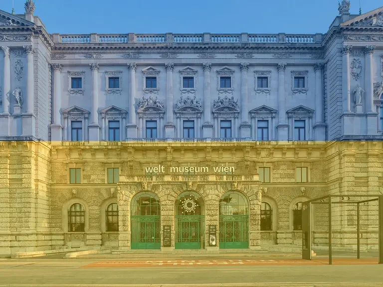 The image shows the exterior of the Weltmuseum Wien, a historic building in Vienna. The façade features ornate architectural details and large windows, with green doors at the entrance. The sky is a soft blue, enhancing the museum's grandeur.