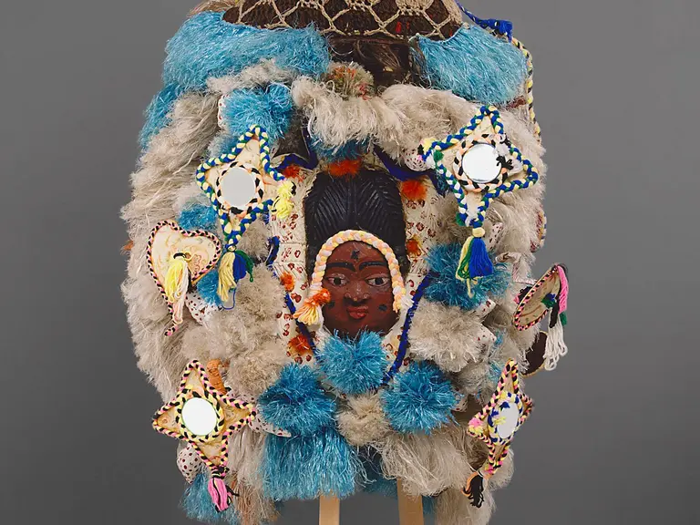 Eine kunstvolle Maske, die aus verschiedenen Materialien gefertigt ist. Sie zeigt mehrere Gesichter, umgeben von bunten Verzierungen und weichen Fellen. Die Farben reichen von Blau bis Erdtönen, und einige Elemente reflektieren das Licht. Die Maske wird auf einem Ständer präsentiert.