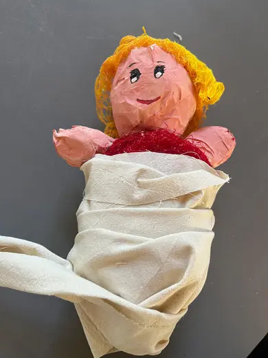 A handmade doll with a pink face and yellow hair is wrapped in a white cloth. The doll has a joyful expression with rosy cheeks and is wearing a red outfit. The background is a simple gray surface.