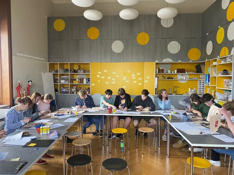 Eine Gruppe von Jugendlichen sitzt an Tischen in einem hellen Raum mit gelben und grauen Wänden. Sie arbeiten an kreativen Projekten, nutzen Papier und Farben. Einige haben Notizen und Materialien vor sich, während sie konzentriert an ihren Aufgaben arbeiten.