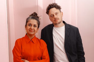 Ein Paar steht vor einer pinken Wand. Die Frau trägt ein rotes Hemd mit hochgeschlossenem Kragen und hat einen Dutt mit Haarschmuck. Der Mann steht neben ihr in einem schwarzen Blazer und einem weißen T-Shirt. Beide lächeln leicht in die Kamera.