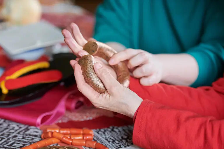 Two hands are shown holding a curved, textured object, while another hand is gently reaching towards it. The background includes various craft materials, suggesting a creative activity.