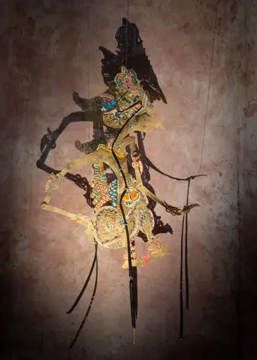 A colorful shadow puppet, intricately decorated with patterns, is suspended against a textured background. The puppet has elongated limbs and is positioned to suggest movement, with its shadow cast dramatically on the wall behind it. This art form highlights traditional storytelling techniques.