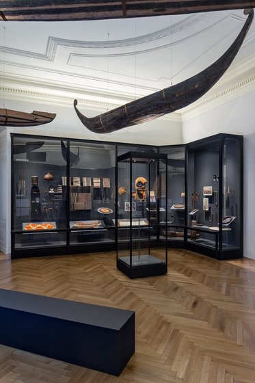 A museum exhibit featuring glass display cases containing various artifacts, including traditional masks, tools, and decorative items. A large canoe is suspended from the ceiling, and a sleek black bench is positioned in the foreground on a wooden floor. The space is well-lit and showcases cultural objects.