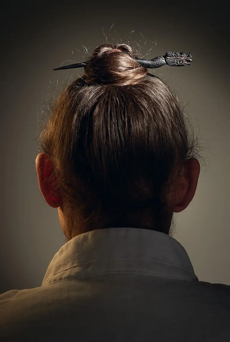 Die Rückansicht einer Person mit langen, dunklen Haaren, die zu einem Dutt hochgesteckt sind. Im Dutt steckt ein dekorativer Haarnadel mit einem einzigartigen Design. Die Person trägt ein helles, schlichtes Oberteil. Der Hintergrund ist dimm und fokussiert auf die Haarfrisur.