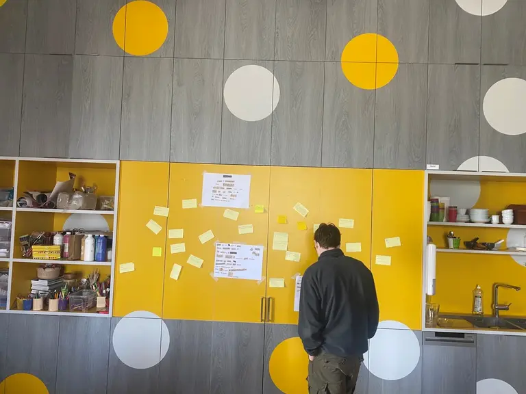 Eine Person steht vor einer gelben Wand mit großen runden, weißen und gelben Punkten. An der Wand kleben zahlreiche Post-its und Zettel mit Informationen. Im Hintergrund sind Küchenschränke mit verschiedenen Utensilien sichtbar.