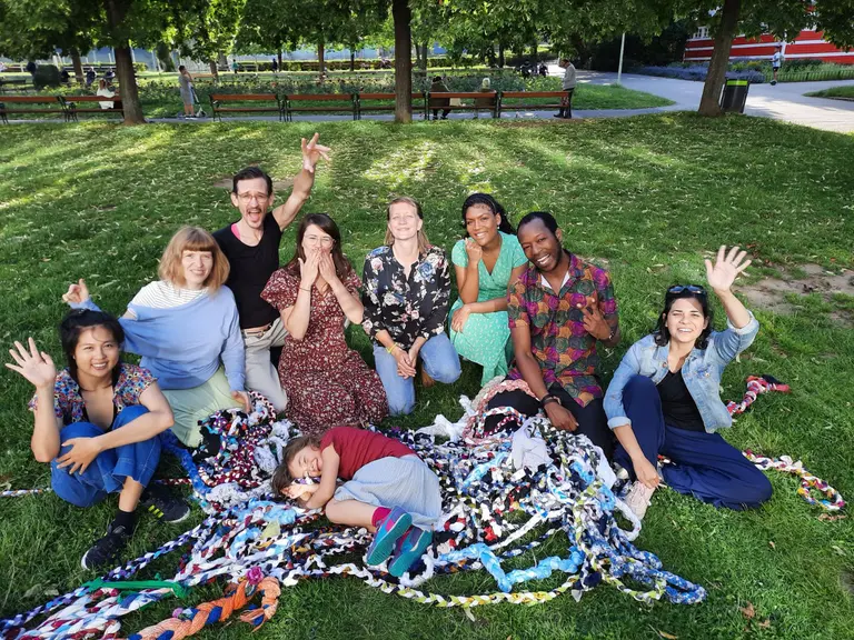 A diverse group of eight people, including adults and a child, sit together on green grass. They are smiling and waving at the camera, with some hands raised in a cheerful manner. Colorful fabric strips are scattered around them, adding a vibrant touch to the scene.