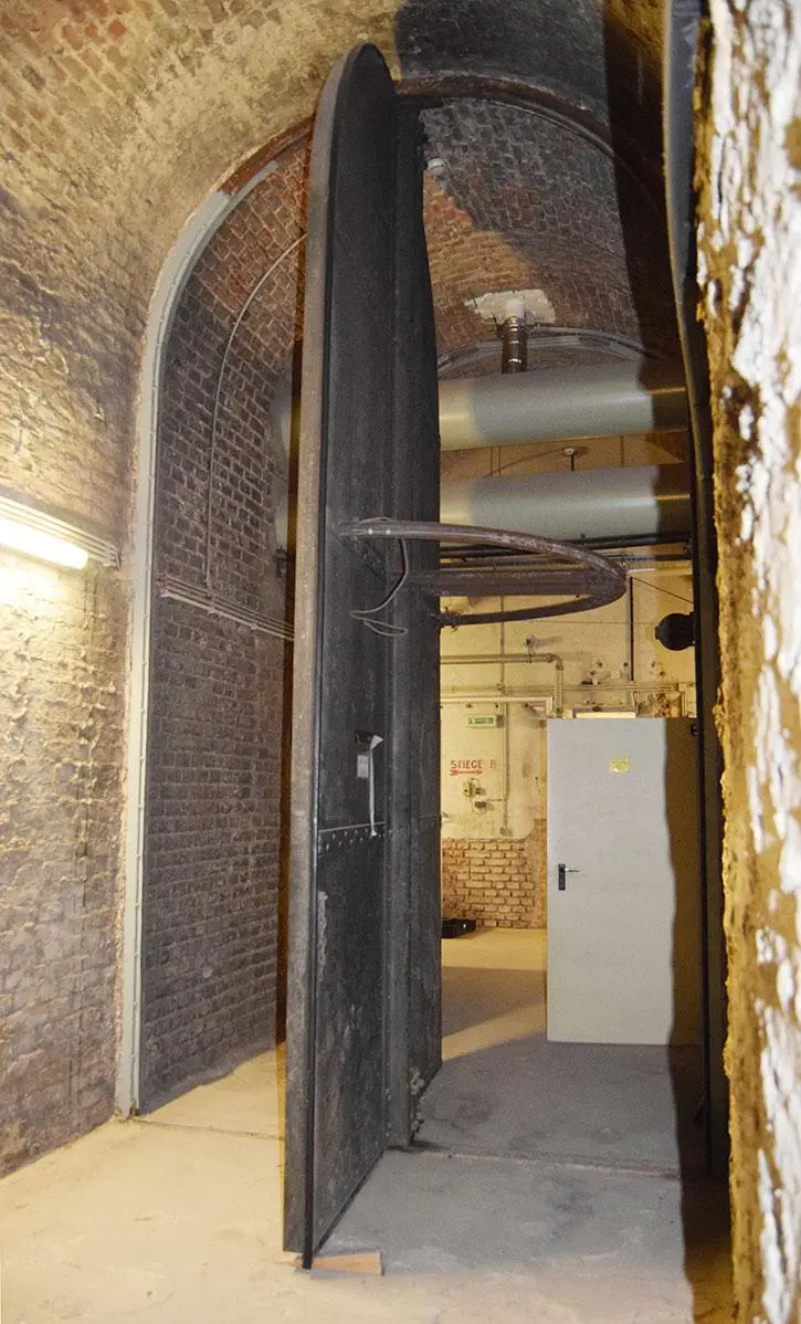 A large, heavy door stands ajar in a dimly lit, industrial space. The walls are made of exposed brick, and pipes are visible in the ceiling. A cabinet can be seen in the background, adding to the utilitarian atmosphere.