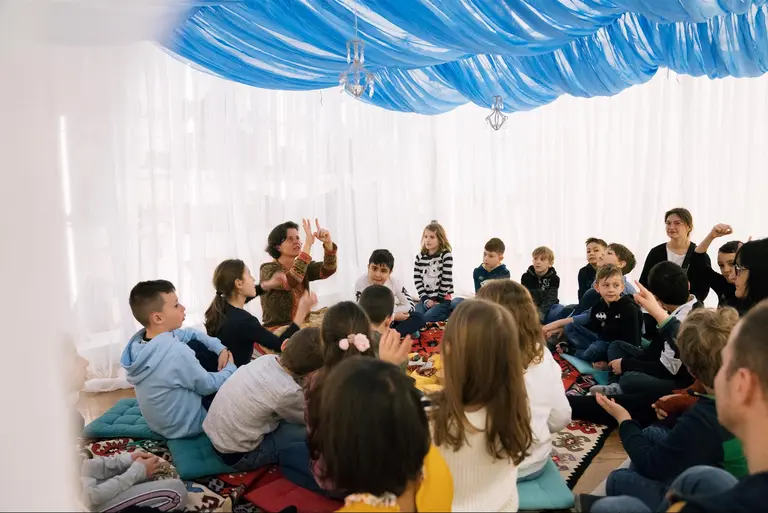 Eine Gruppe von Kindern und Erwachsenen sitzt im Kreis in einem hellen Raum mit buntem Teppich. Eine Person in der Mitte spricht, während die anderen aufmerksam zuhören und einige ihre Hände heben, als würden sie Fragen stellen oder interagieren. Der Raum ist mit blauer Decke dekoriert.