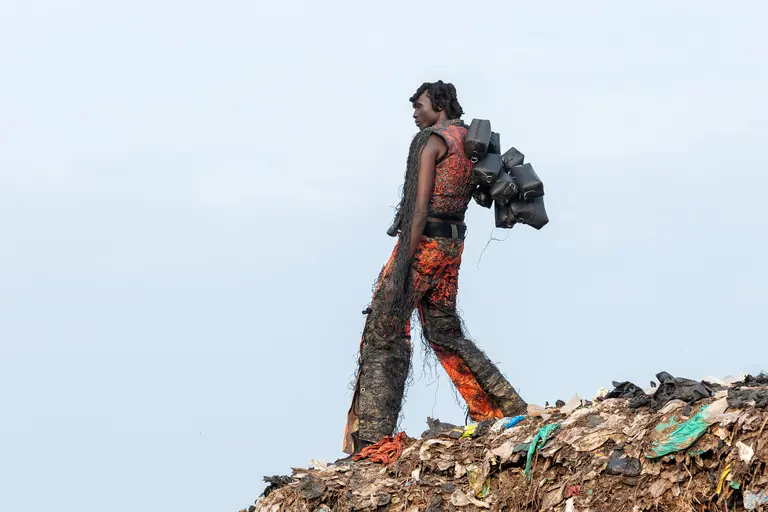 Ein Mensch mit kreativem Outfit, das aus Müll und Recyclingmaterialien besteht, geht über einen Müllhaufen. Der Hintergrund zeigt einen hellen Himmel. Die Szene symbolisiert Umweltbewusstsein und die Auswirkungen von Abfall auf die Gesellschaft.