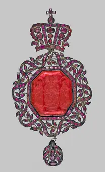 A decorative medallion featuring a central red octagonal gemstone surrounded by intricate metalwork with floral designs. At the top, a crown is adorned with small gems, and at the bottom hangs a rounded ornament. The overall design is ornate and regal.