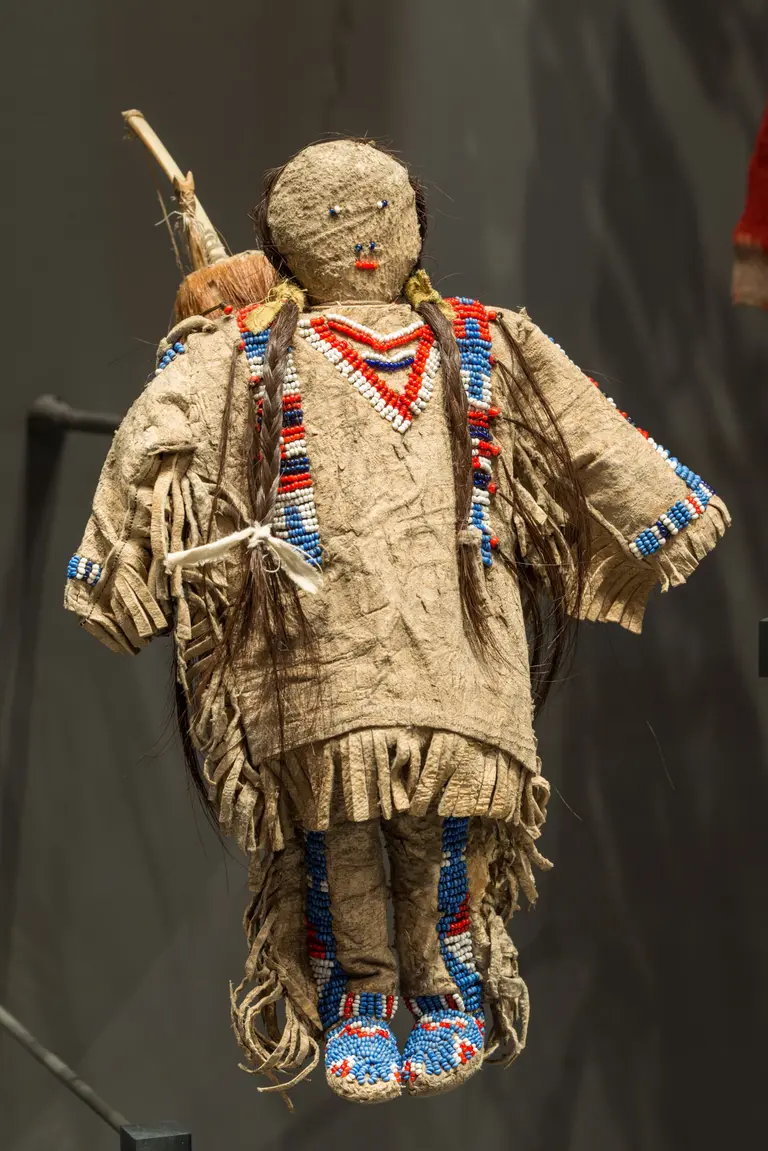 Eine traditionelle Puppe, gefertigt aus Stoff und Naturmaterialien. Sie trägt ein buntes Gewand mit Fransen und ist mit Perlen verziert. Ihr Gesicht ist schlicht gehalten, und sie hat lange Haare. Die Puppe repräsentiert möglicherweise kulturelle Traditionen und Handwerkskunst.