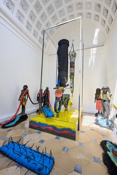 Eine beeindruckende Installation in einem hellen Raum. Im Mittelpunkt steht ein schwarzes, fahrbares Objekt, umgeben von bunten Skulpturen und Textilien, die menschliche Figuren darstellen. Der Boden ist mit hellen Fliesen gestaltet, die das Gesamtbild der Kunstinstallation ergänzen.