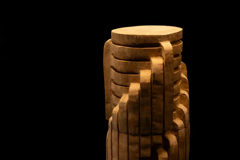 Eine künstlerische Holzskulptur mit gestapelten, geschwungenen Formen und einem glatten, flachen oberen Teil. Die Skulptur hat eine natürliche Holzfarbe und steht vor einem schwarzen Hintergrund, der den Fokus auf die Details der Holzstruktur lenkt.