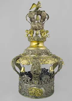 A decorative crown made of metal, featuring intricate designs and embellishments. The crown has a golden top adorned with a bird, surrounded by lion figures. The lower portion includes floral patterns and is finished in silver and gold tones, highlighting its ornate craftsmanship.