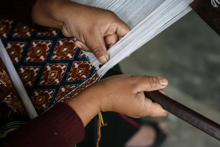 Eine Person arbeitet an einem Webstuhl, ihre Hände halten Fäden und gestalten ein geometrisches Muster. Die Farben sind lebhaft und vielfältig. Der Fokus liegt auf dem Handwerk und der Kunst des Webens.