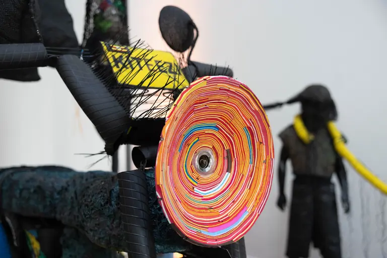 Ein kunstvolles Objekt mit buntem, spiralförmigem Rad aus verschiedenen Materialien. Im Hintergrund ist eine figurative Skulptur zu sehen, die aus Stoff besteht und eine menschliche Gestalt darstellt. Die gesamte Szene wirkt kreativ und modern.