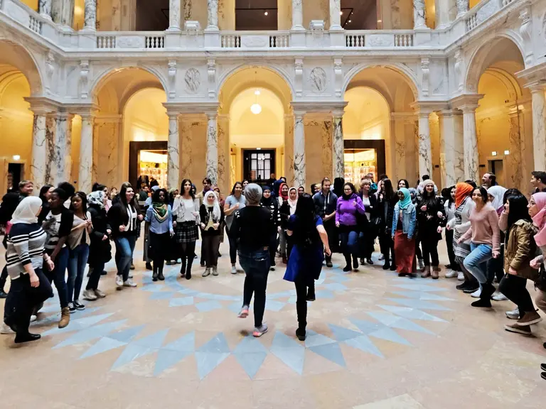 Eine große Gruppe von Menschen versammelt sich in einer weitläufigen, architektonisch beeindruckenden Halle mit hohen Decken und Säulen. Der Boden hat ein auffälliges geometrisches Muster. Die Atmosphäre wirkt lebhaft und dynamisch.