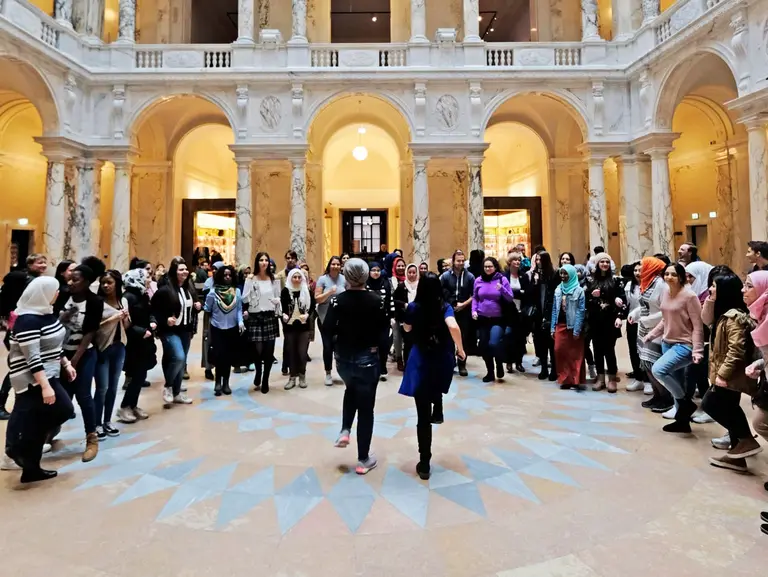 Eine große Gruppe von Menschen versammelt sich in einer weitläufigen, architektonisch beeindruckenden Halle mit hohen Decken und Säulen. Der Boden hat ein auffälliges geometrisches Muster. Die Atmosphäre wirkt lebhaft und dynamisch.