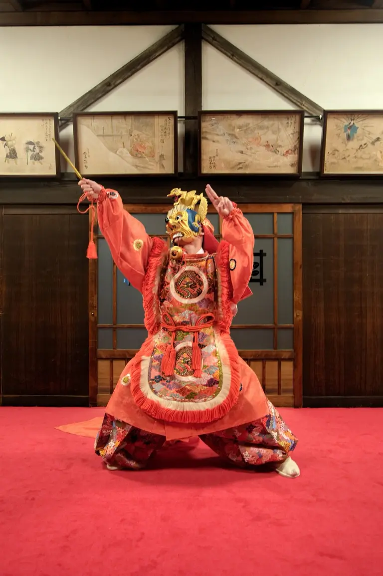 A performer in elaborate traditional clothing striking a dramatic pose on a red carpeted stage. The costume features bright colors and intricate designs, showcasing cultural patterns. The background includes wooden walls with framed artworks, enhancing the theatrical atmosphere.