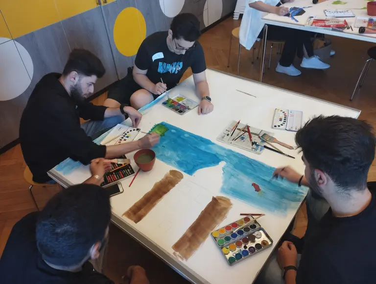 Fünf junge Männer sitzen an einem Tisch und malen gemeinsam auf ein großes Blatt Papier. Sie verwenden Wasserfarben und Pinsel, um eine Landschaft zu gestalten. Farbpaletten und Malutensilien sind um sie herum verteilt. Der Hintergrund ist hell und modern gestaltet.