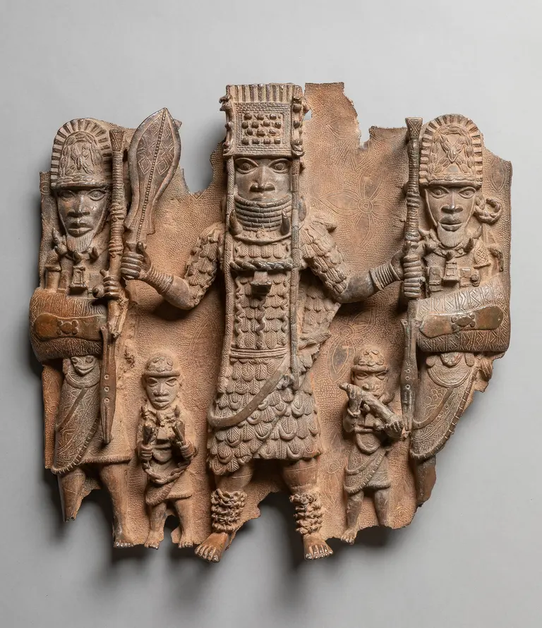 Ein reliefartiges Kunstwerk, das einen Herrscher zeigt, umgeben von zwei Wächtern und einem kleineren Figuren. Der Herrscher trägt aufwendige Kleidung und eine beeindruckende Kopfbedeckung, während die Wächter Schilde und Speere halten. Der Hintergrund ist strukturiert und unregelmäßig.
