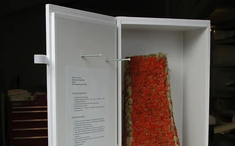 A white display cabinet is partially open, revealing a brightly colored, textured object resembling a woven textile on the interior. A piece of paper with text is attached to the side of the cabinet, providing information about the displayed item.