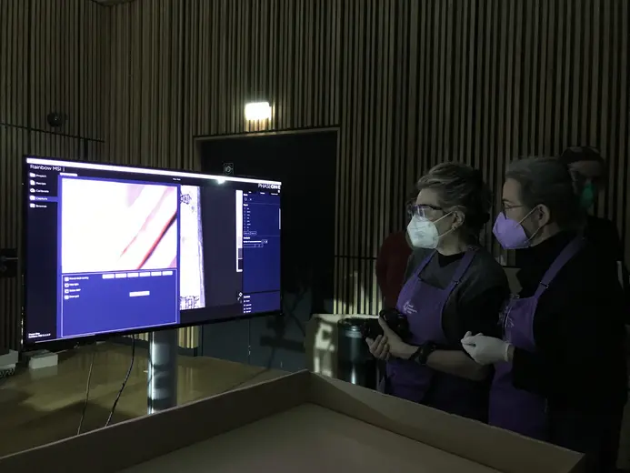 Zwei Personen in Schürzen stehen vor einem Monitor, auf dem ein Bild gezeigt wird. Beide tragen Schutzmasken und schauen interessiert auf den Bildschirm, der Details einer mikroskopischen Untersuchung zu zeigen scheint. Die Umgebung wirkt modern und gut beleuchtet.