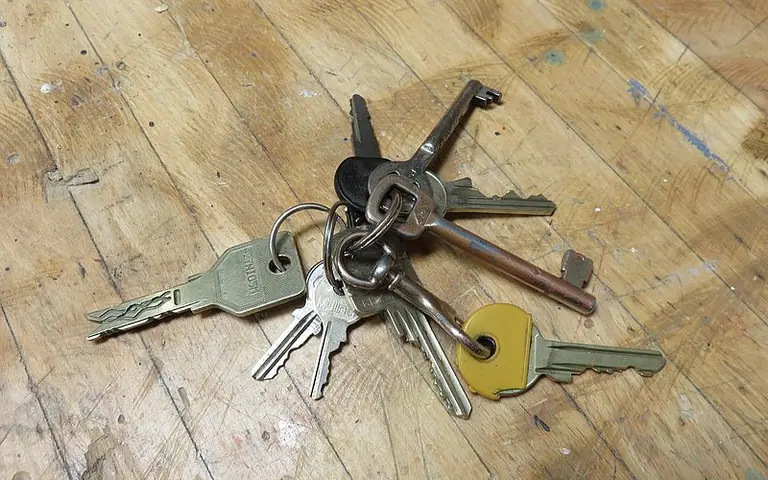 Eine Ansammlung von Schlüsseln liegt auf einem Holzuntergrund. Die Schlüssel sind in verschiedenen Formen und Farben, darunter ein gelber Schlüssel. Sie sind an einem Schlüsselring befestigt, der im Mittelpunkt der Anordnung steht.