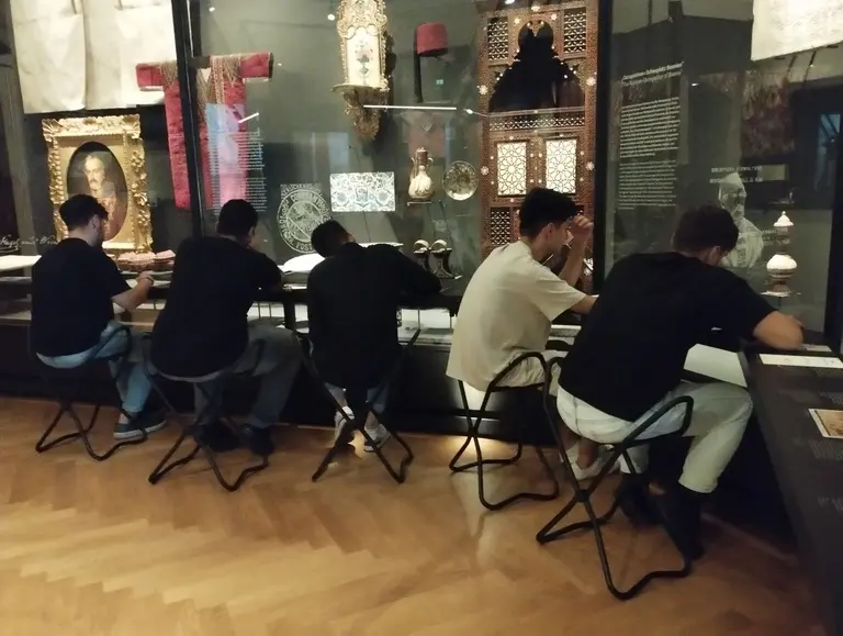 Fünf Personen sitzen an einem Tisch in einem Ausstellungsraum. Sie beschäftigen sich mit den dort präsentierten Kunstwerken und Ausstellungsstücken. Im Hintergrund sind verschiedene historische Objekte und Informationen zu sehen.