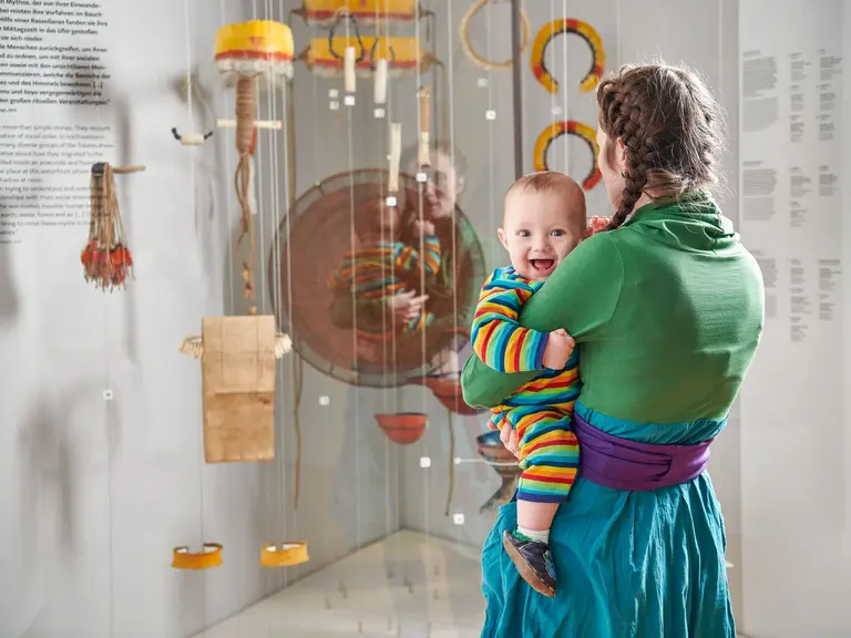 Eine Frau trägt ein lächelndes Baby in einem bunten Streifenanzug. Im Hintergrund sind interessante Objekte in Vitrinen ausgestellt. Die Szene spielt in einem Museum, das kulturelle Exponate zeigt.