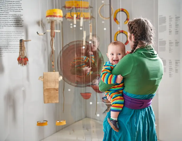 Eine Frau trägt ein lächelndes Baby in einem bunten Streifenanzug. Im Hintergrund sind interessante Objekte in Vitrinen ausgestellt. Die Szene spielt in einem Museum, das kulturelle Exponate zeigt.