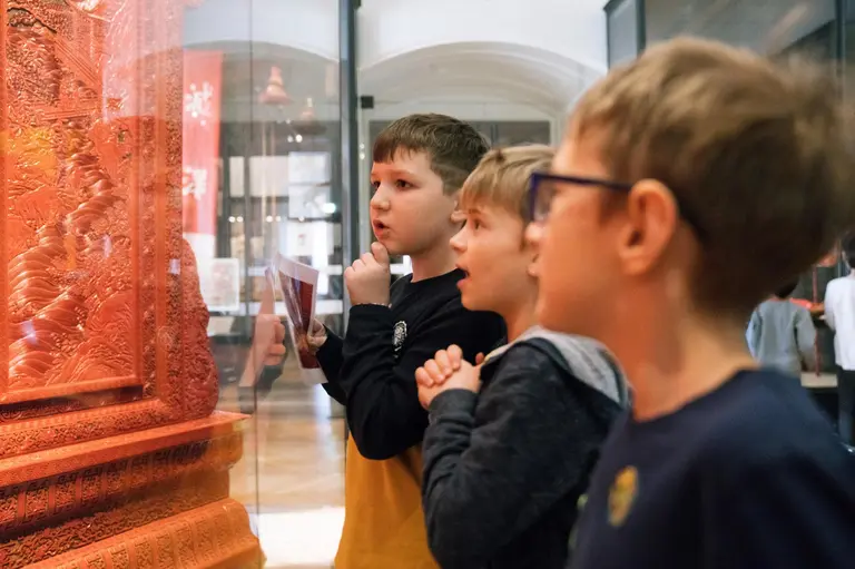 Drei Kinder stehen vor einem Ausstellungsstück in einem Museum. Sie schauen fasziniert und nachdenklich auf die Details des Kunstwerks, das in einem Glaskasten präsentiert wird. Die Atmosphäre ist neugierig und erforschend.