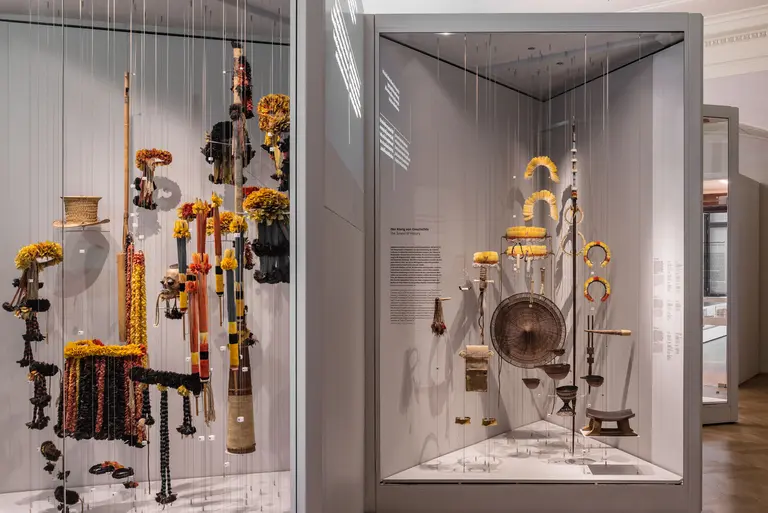 Das Bild zeigt eine Ausstellung mit traditionellen Kunstobjekten, die in Vitrinen präsentiert sind. Die Objekte sind farbenfroh dekoriert, darunter Figuren und Musikinstrumente. Es gibt eine informative Beschilderung neben den Ausstellungsstücken, die deren Ursprung und Bedeutung erklärt.