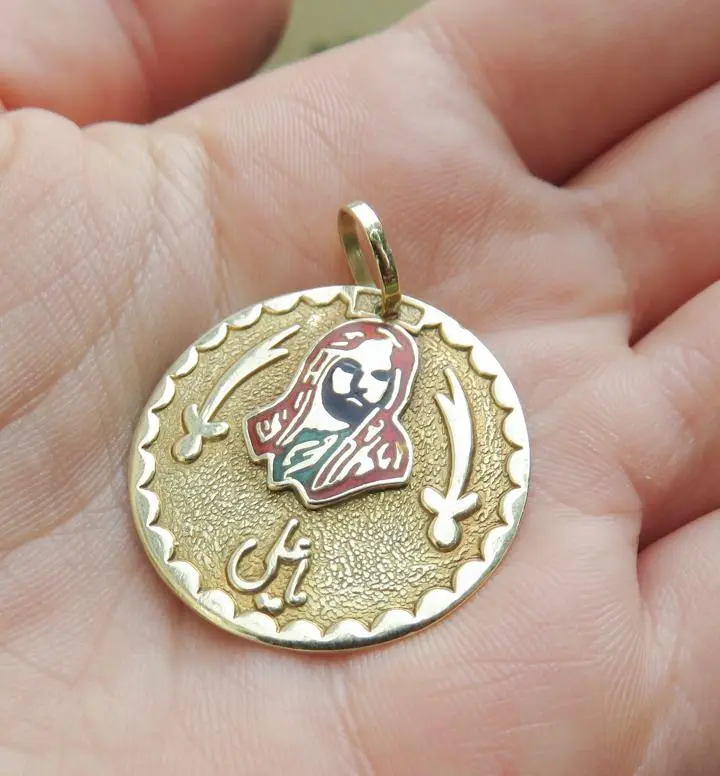 Ein runder, goldfarbener Anhänger liegt in einer Hand. Auf der Vorderseite ist ein stilisiertes Porträt eines Mannes und zwei gekreuzte Schwerter abgebildet. Unter dem Porträt sind arabische Schriftzeichen zu sehen. Der Anhänger hat eine glänzende Oberfläche.